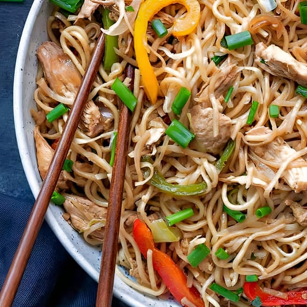 Close-up of Indian chicken noodles with colorful bell peppers, green onions, and chopsticks.
