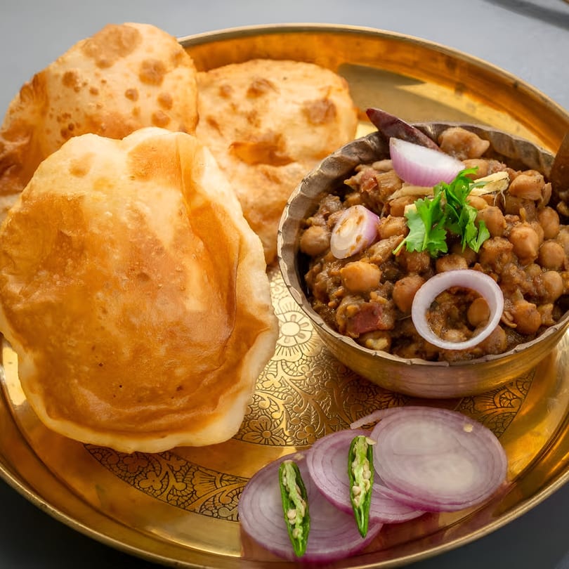 Golden puri bread and chana masala with red onion and green chili on a brass plate.