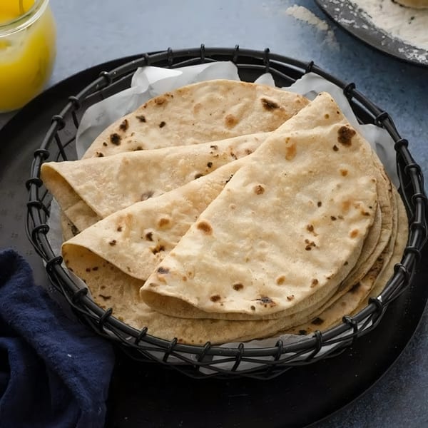 A black wire basket filled with folded, warm Indian rotis, with a jar of ghee nearby.