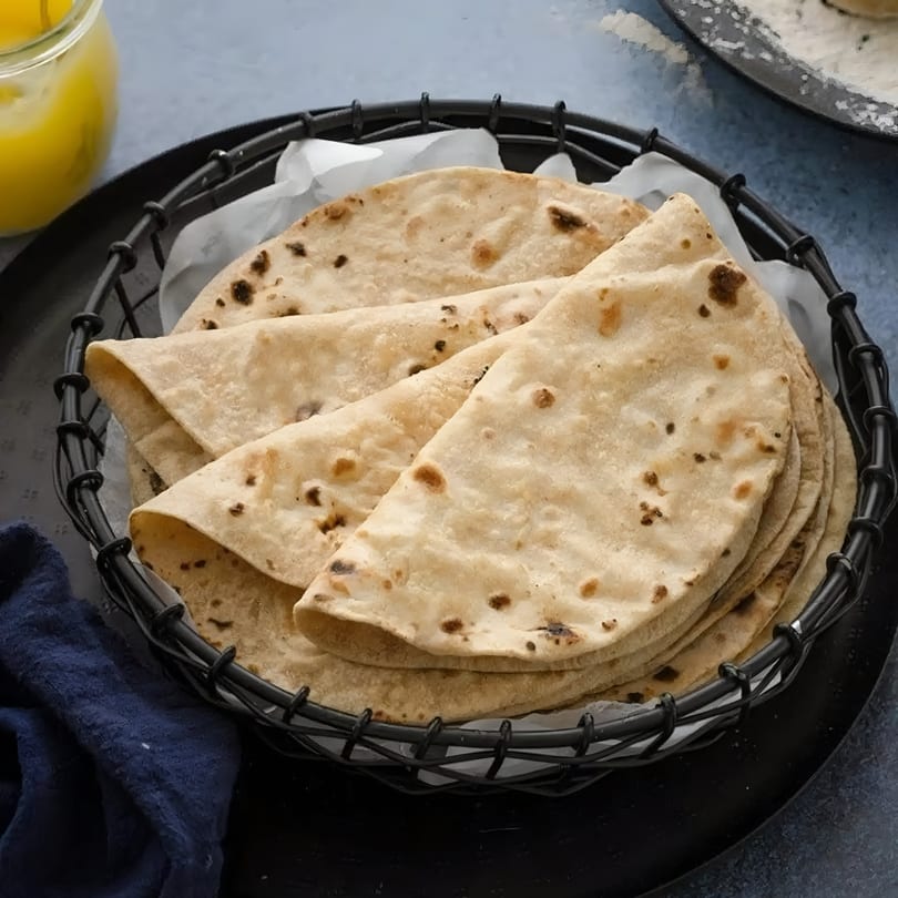 A black wire basket filled with folded, warm Indian rotis, with a jar of ghee nearby.